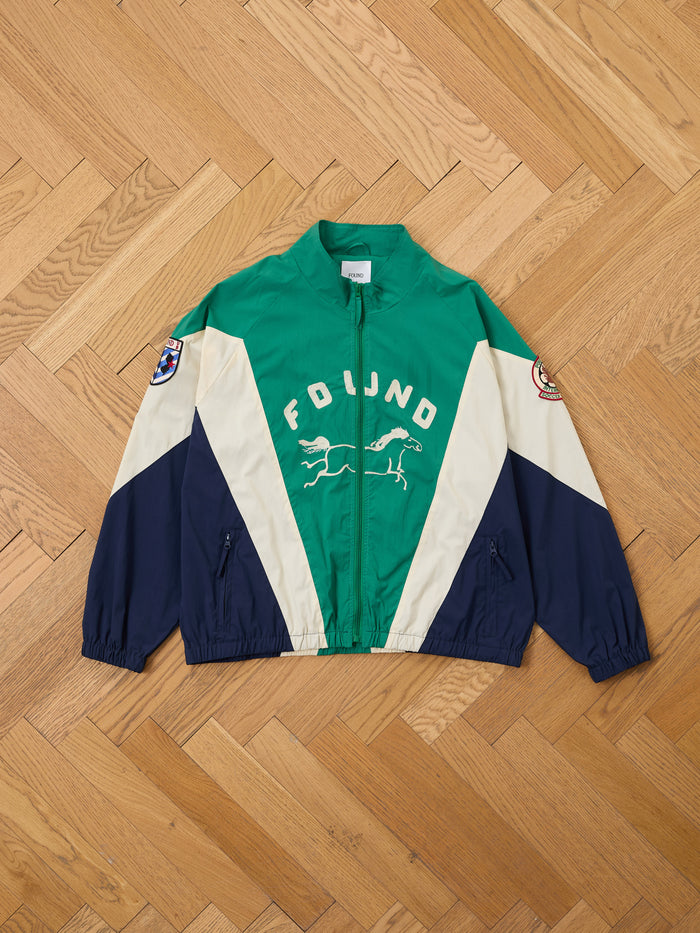 The Crestline Sport Zip Jacket by FOUND features green, navy, and cream panels, "FOUND" text, and two running horses on the front. Styled on a wooden parquet floor, this lightweight windbreaker channels vintage sportswear vibes.