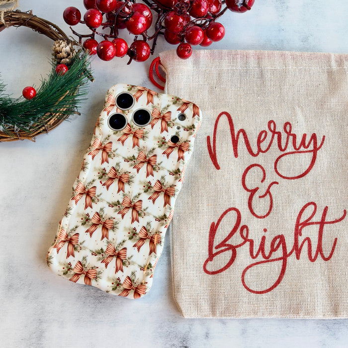 Deck The Halls With Bows & Holly iPhone case lying flat beside a Merry & Bright linen bag, a pine wreath and red berries