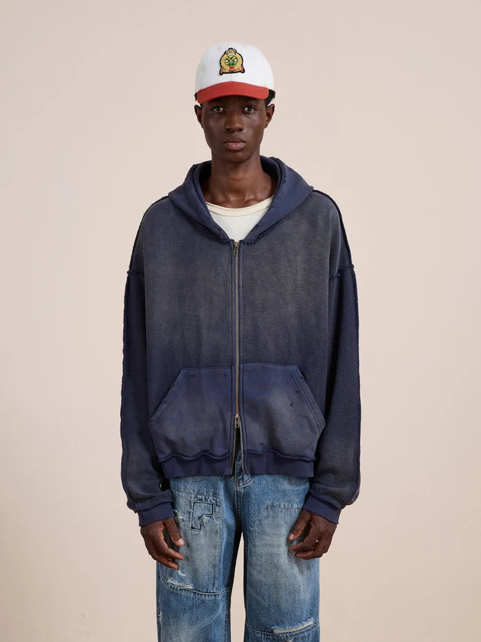 A person in a white and red cap, FOUND Deconstructed Terry Zip Up Hoodie, and distressed blue jeans stands against a plain beige background.