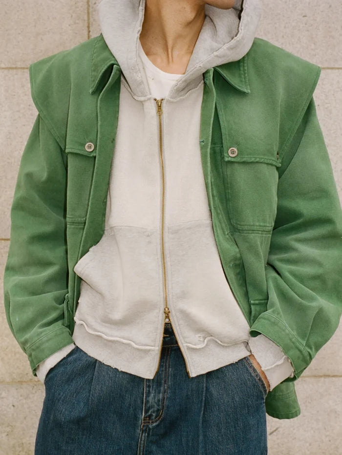 A person wears the FOUND Deconstructed Terry Zip Up Hoodie over a white T-shirt, paired with blue jeans, and stands against a light stone wall.