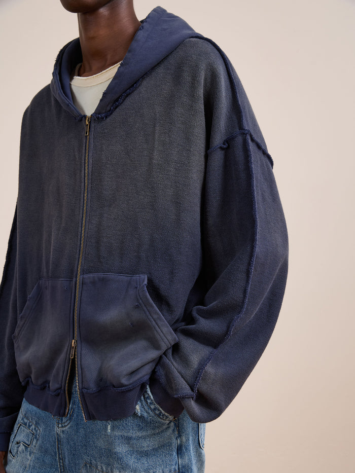 A person in a faded navy FOUND Deconstructed Terry Zip Up Hoodie over a beige t-shirt and blue jeans stands against a plain light background.