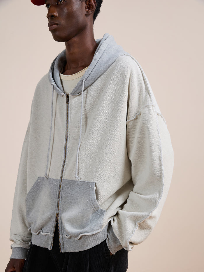 A person wearing FOUND's Deconstructed Terry Zip Up Hoodie in light gray, featuring a front pocket and drawstrings, layered over a cream shirt with a red cap, standing against a plain background.