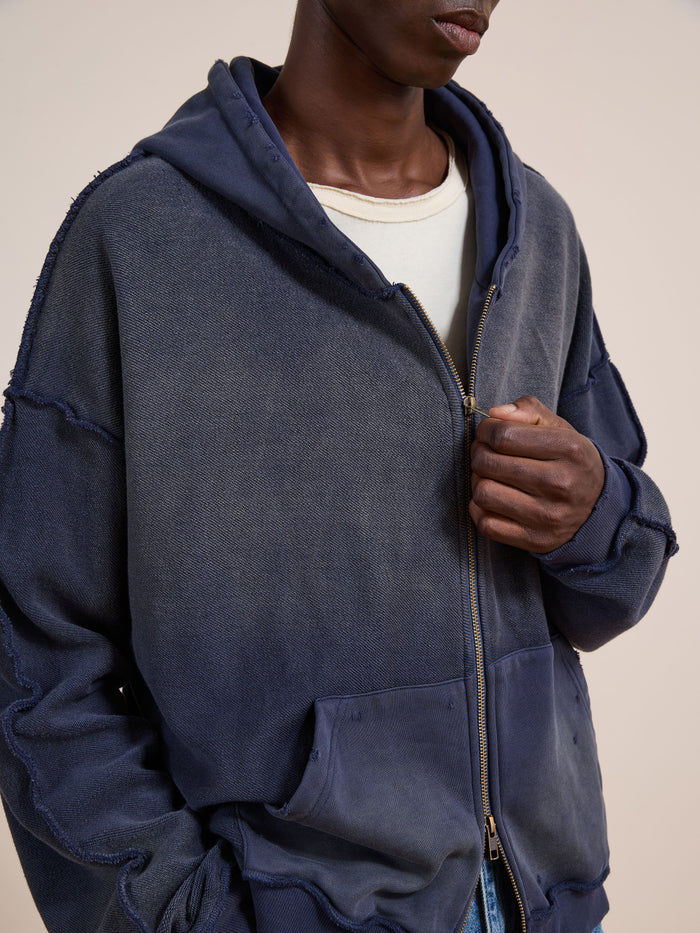 A person wears the FOUND Deconstructed Terry Zip Up Hoodie in faded navy blue over a white t-shirt, holding the zipper with one hand.