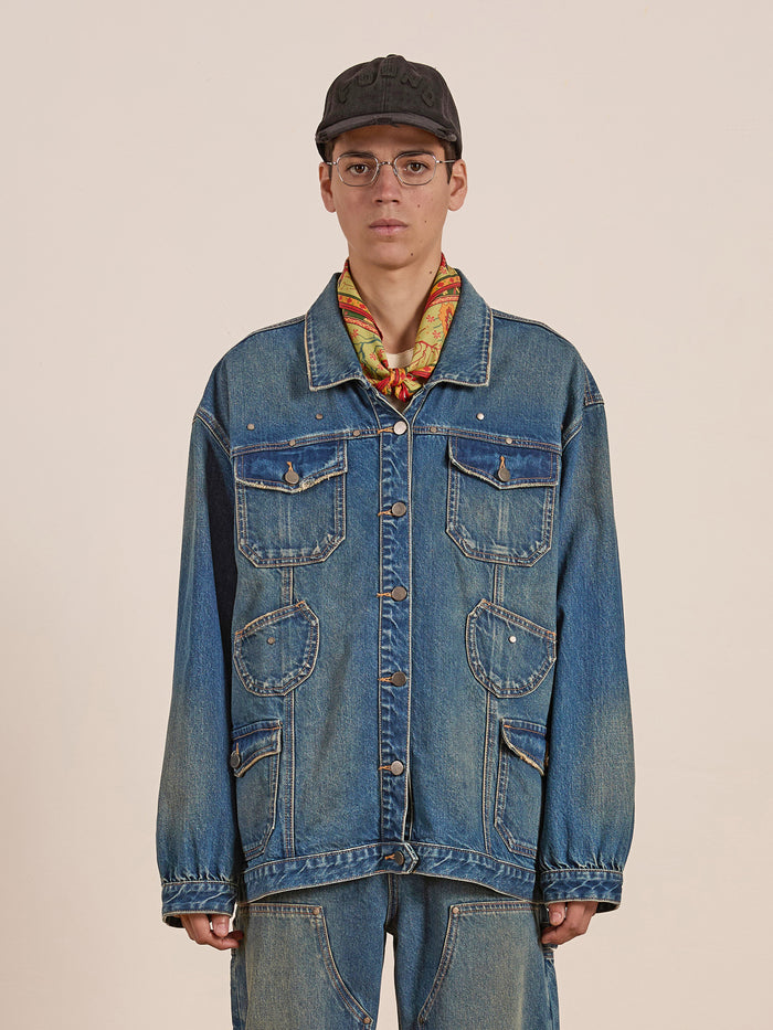 A person stands confidently against a plain background wearing FOUND’s Denim Carpenter Jacket with matching relaxed-fit jeans, accessorized by a black cap, glasses, and a colorful scarf.
