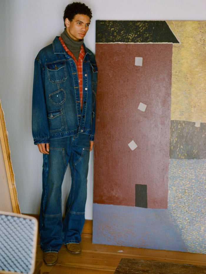 A person stands by a wall in FOUND's Denim Carpenter Jacket and jeans, with a sweater underneath. This unisex relaxed-fit outfit is complemented by an abstract painting against the wall, embodying the perfect workwear staple for any creative setting.
