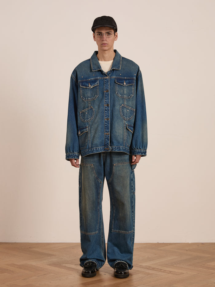 A person in an oversized denim jacket, wearing FOUND's Denim Carpenter Jeans with a vintage-inspired wash, paired with a black cap and glasses, stands on a wooden floor against a plain background.