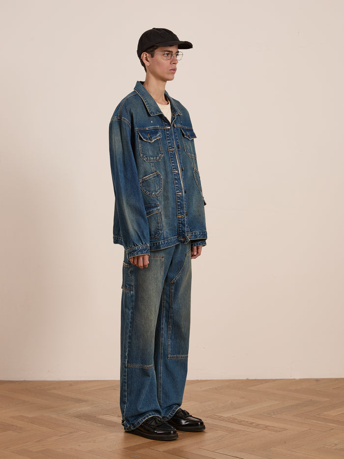 Wearing a Denim Carpenter Jacket by FOUND, this individual in jeans, glasses, and a black cap stands on a wooden floor against a beige backdrop, showcasing the timeless appeal of workwear.