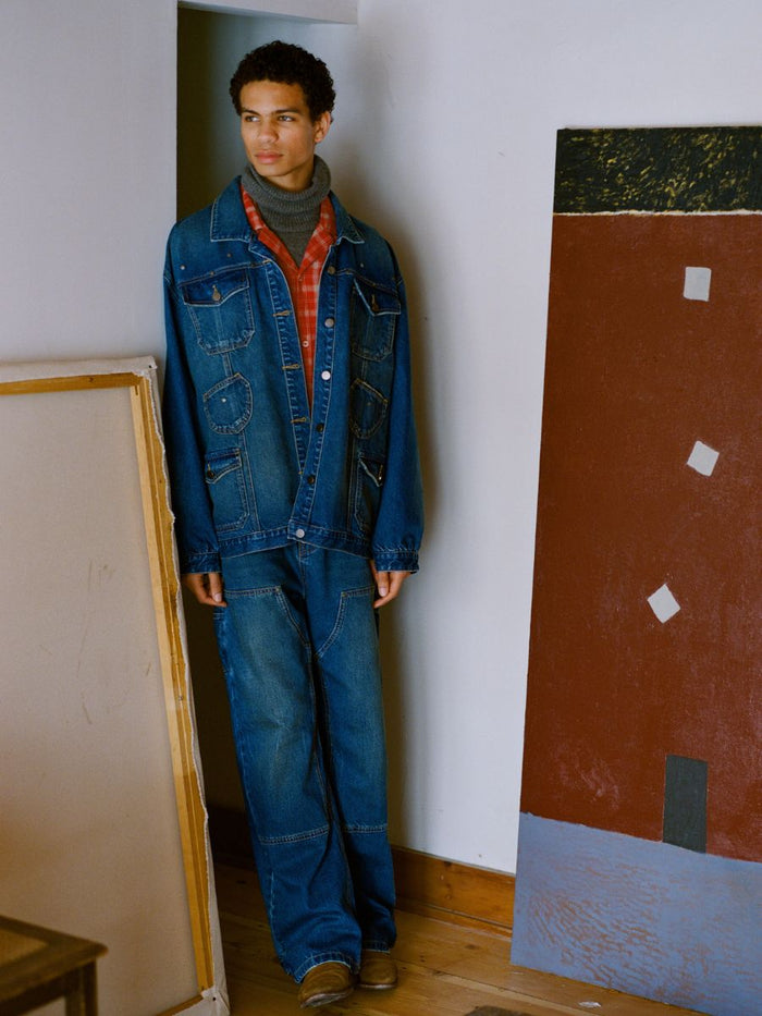 A person in a denim jacket and FOUND's Denim Carpenter Jeans featuring a vintage-inspired wash stands by a wall adorned with framed artworks in the room.
