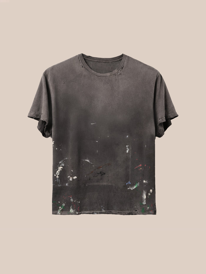 The FOUND Distressed Paint Tee, a unisex relaxed fit shirt in dark gray with faded paint stains and subtle distressing, is displayed against a neutral beige background.