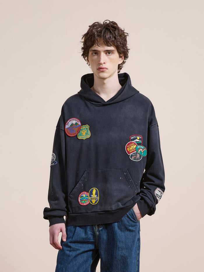 A young person in the FOUND Distressed Patch Hoodie—a unisex relaxed fit with colorful embroidered patches—paired with blue jeans stands against a beige background, gazing at the camera.