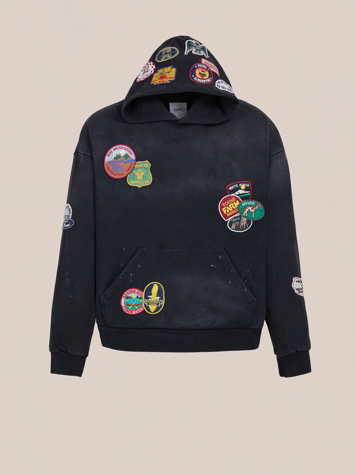 The FOUND Distressed Patch Hoodie in vintage washed black showcases multiple colorful embroidered patches on the front, hood, sleeves, and pocket, set against a plain light background.
