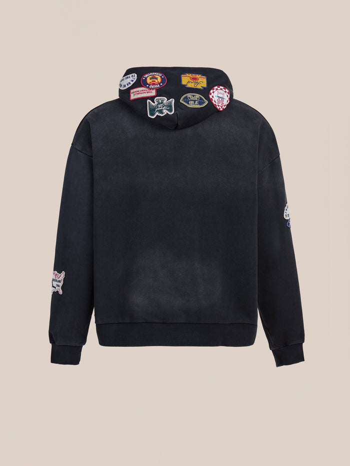 The FOUND Distressed Patch Hoodie is a unisex relaxed fit black sweatshirt shown from the back, with colorful embroidered patches on the hood and sleeves, set against a beige background.