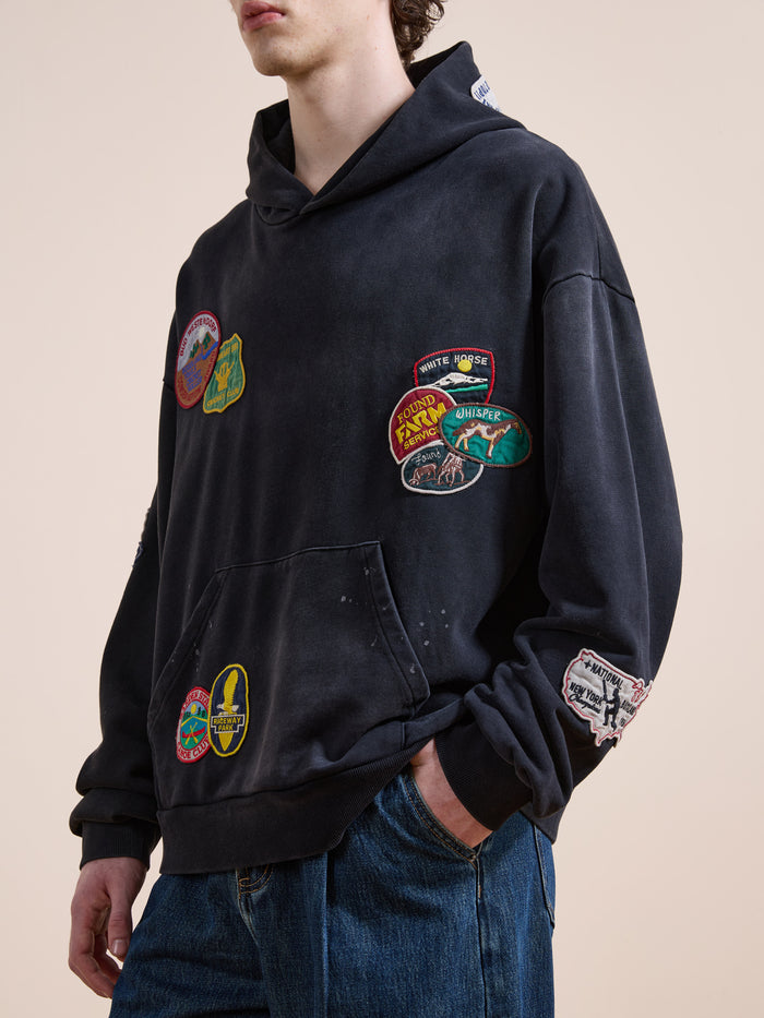 A person wears the FOUND Distressed Patch Hoodie, a vintage washed black unisex relaxed fit top with colorful patches, paired with blue jeans, standing against a plain background.