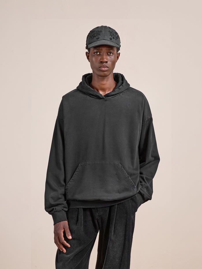 A person wearing the FOUND Distressed Pullover Hoodie in washed black, paired with black pants and a black cap, stands against a plain beige background.