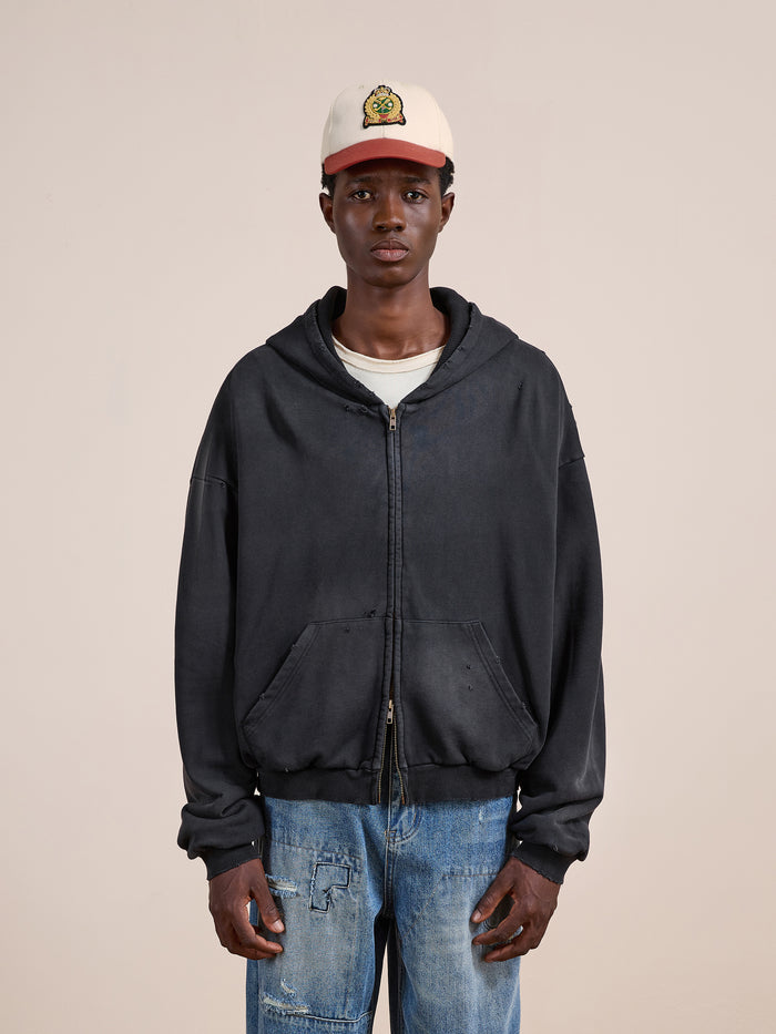 A person stands against a beige background wearing the FOUND Distressed Thermal Lined Zip Up Hoodie in black, a white t-shirt, blue jeans, and a white cap featuring a cartoon graphic.