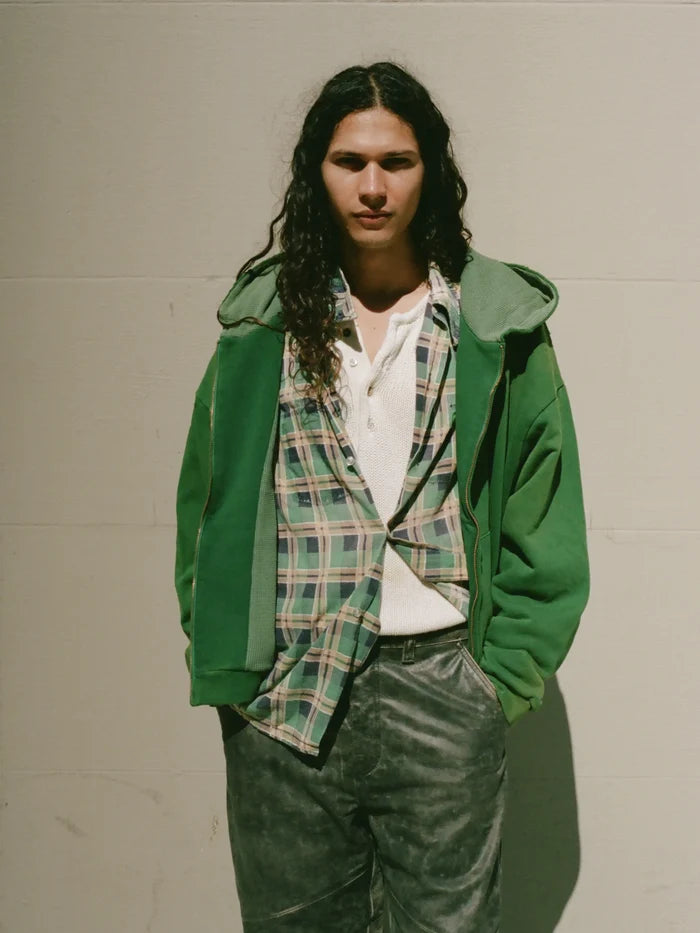 A person with long curly hair stands against a plain wall, wearing a green FOUND Distressed Thermal Lined Zip Up Hoodie over a plaid shirt and white top, paired with grey pants and has hands casually tucked in pockets.