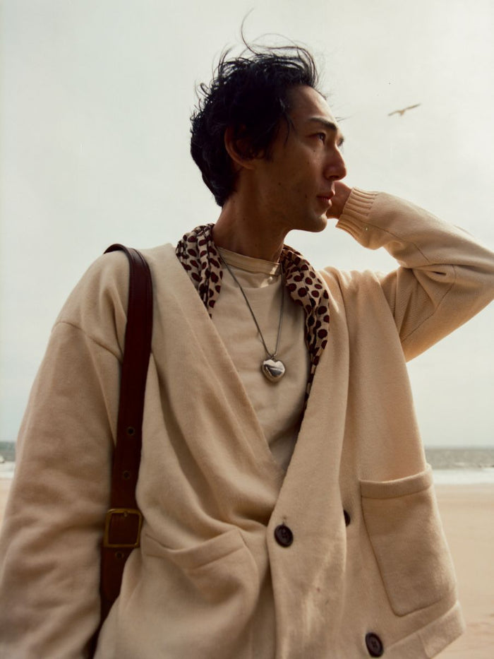 Wearing FOUND's Double Breasted Knit Cardigan, a person stands on the beach, gazing into the distance with one hand touching their head—a relaxed unisex look blending effortless tailoring and knitwear.