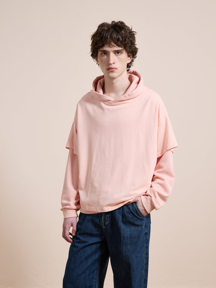 A young man with wavy brown hair wears the Found Double Layer Hoodie, a unisex relaxed fit in pink French terry cotton, paired with blue jeans. He stands against a plain beige background with one hand in his pocket.