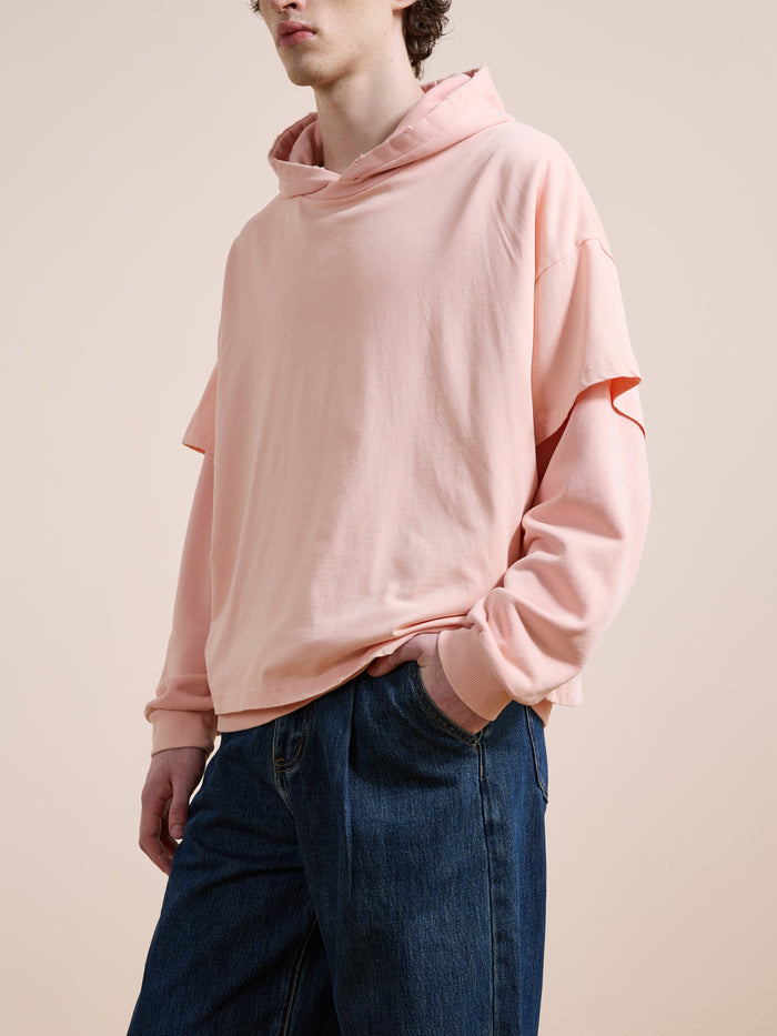 A person in Found’s Double Layer Hoodie—unisex relaxed fit, light pink French terry cotton—pairs it with blue jeans and stands against a plain beige background, one hand in their pocket.