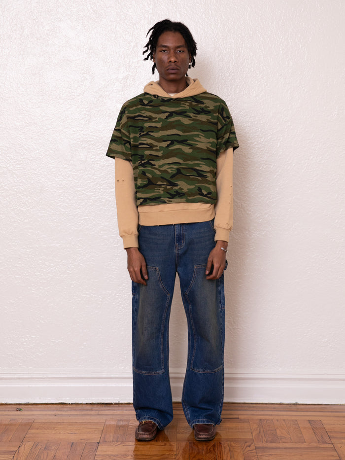 A person stands against a white wall wearing a camouflage shirt over the Double Layer Hoodie by FOUND in beige, paired with vintage-look blue jeans and brown shoes.
