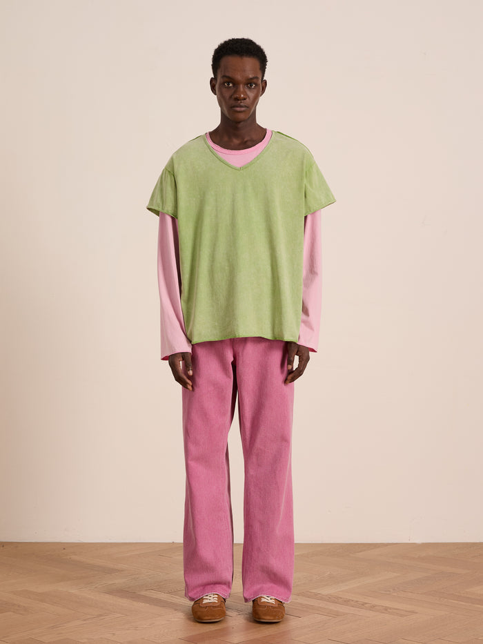 A person stands on a wooden floor wearing FOUND's green Double Layer LS Tee over a pink long sleeve tee, with pink pants and brown shoes, set against a plain beige background.