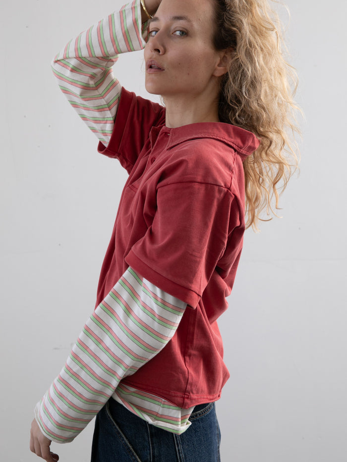 A person with curly blonde hair wears the FOUND Double Layer Stripe Polo in red over a striped long-sleeve top and blue jeans, creating a vintage athletic look against a plain light background.