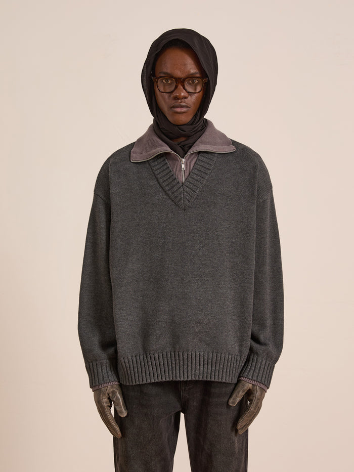A person in glasses and a headscarf wears the FOUND Double Layer Sweater, an oversized grey wool blend, with gloves and dark pants against a plain beige background—ideal for cold-weather layering.