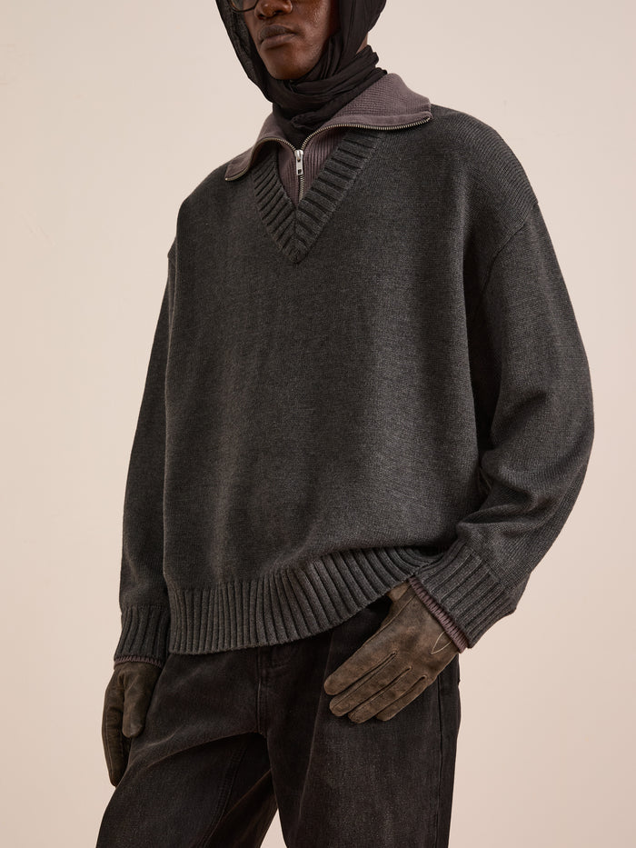 A person showcases practical cold-weather layering in the FOUND Double Layer Sweater—a dark grey wool blend—over a zip-collared shirt, paired with dark jeans, brown gloves, and a hooded head covering, standing against a plain background.