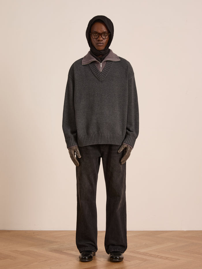 A person stands against a plain wall wearing FOUND’s Double Layer Sweater—a wool blend gray pullover over a dark hoodie, paired with wide-leg black pants, gloves, glasses, and black shoes—ideal for cold-weather layering.