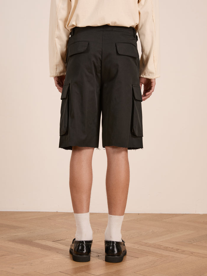 A person stands on a wooden floor, facing away, wearing a beige long-sleeve shirt and Found Elbas Cargo Shorts in black with dual cargo pockets and adjustable waist tabs, paired with white socks and black shoes.