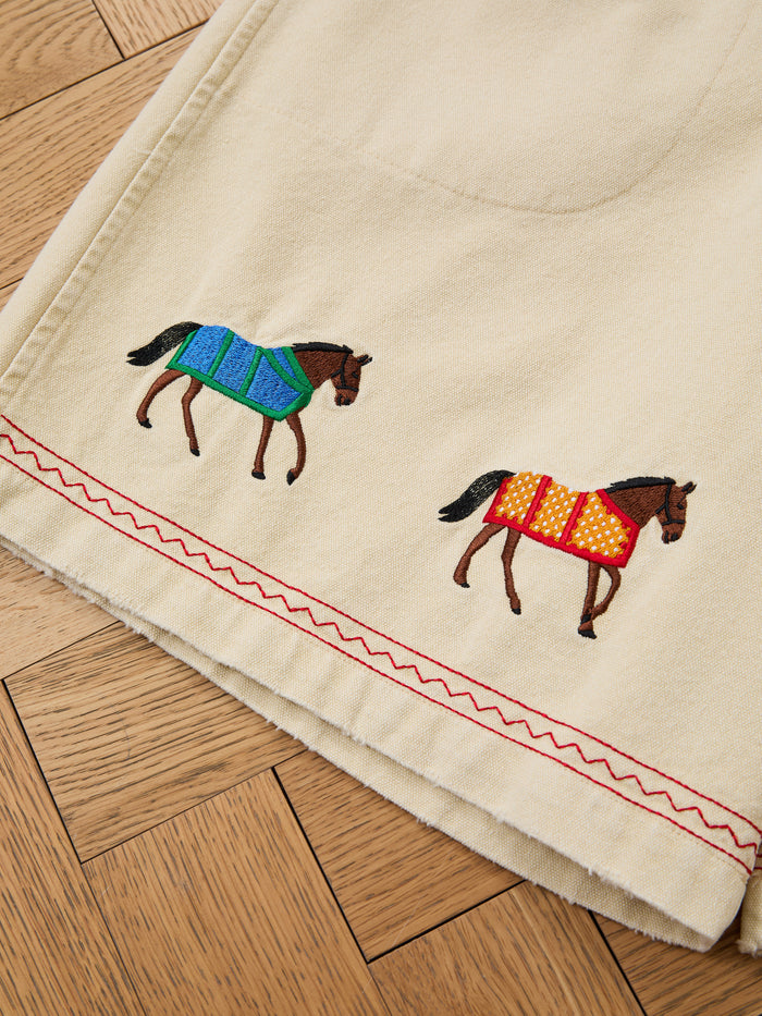 Profound Embroidered Horse Shorts in beige cotton twill, featuring red zigzag stitching and two embroidered horses—one with a blue blanket, the other with a yellow blanket with red polka dots—shown on a wooden floor.