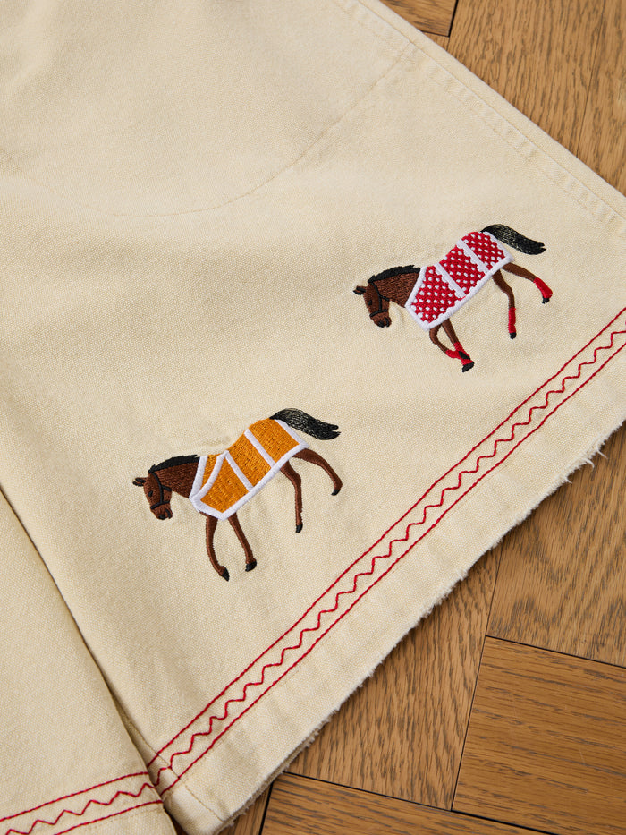 Profound's Embroidered Horse Shorts feature beige cotton twill, red zigzag stitching, and playful horse embroidery—one in a red patterned blanket, the other in yellow—offering unique charm to your wardrobe.