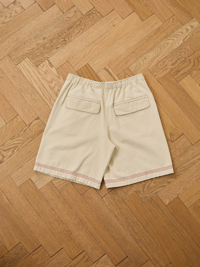 Profound Embroidered Horse Shorts in beige cotton twill with elastic waistband, two back flap pockets, and red stitching detail along the hem, displayed on a wooden herringbone floor.
