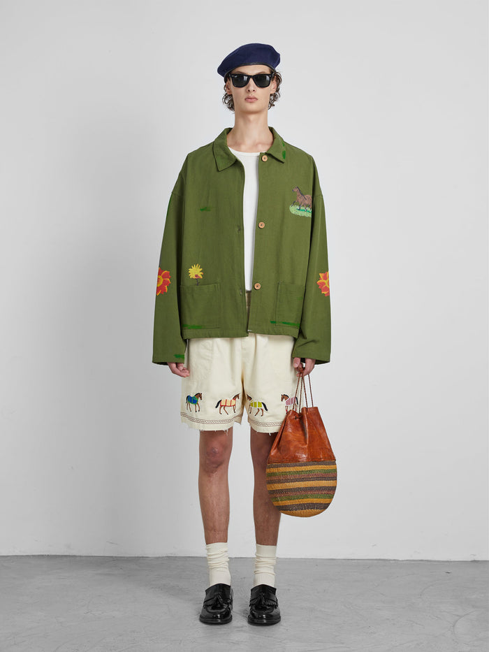 A person wearing a green jacket, the Profound Embroidered Horse Shorts in beige cotton twill, black shoes, and a beret holds a striped brown bag.