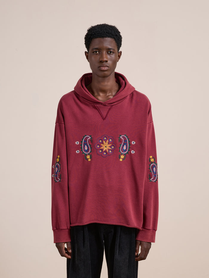A person wearing the FOUND Embroidered Kantha Paisley Hoodie—a loose, unisex relaxed fit hoodie adorned with colorful Kantha embroidery on the front and sleeves—stands against a plain beige background.