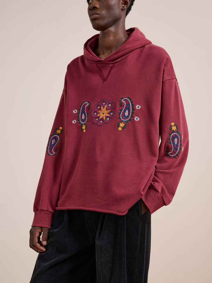 A person wears the FOUND Embroidered Kantha Paisley Hoodie, a unisex relaxed-fit maroon cotton fleece with colorful Kantha-inspired paisley embroidery, paired with dark pants against a plain beige background.