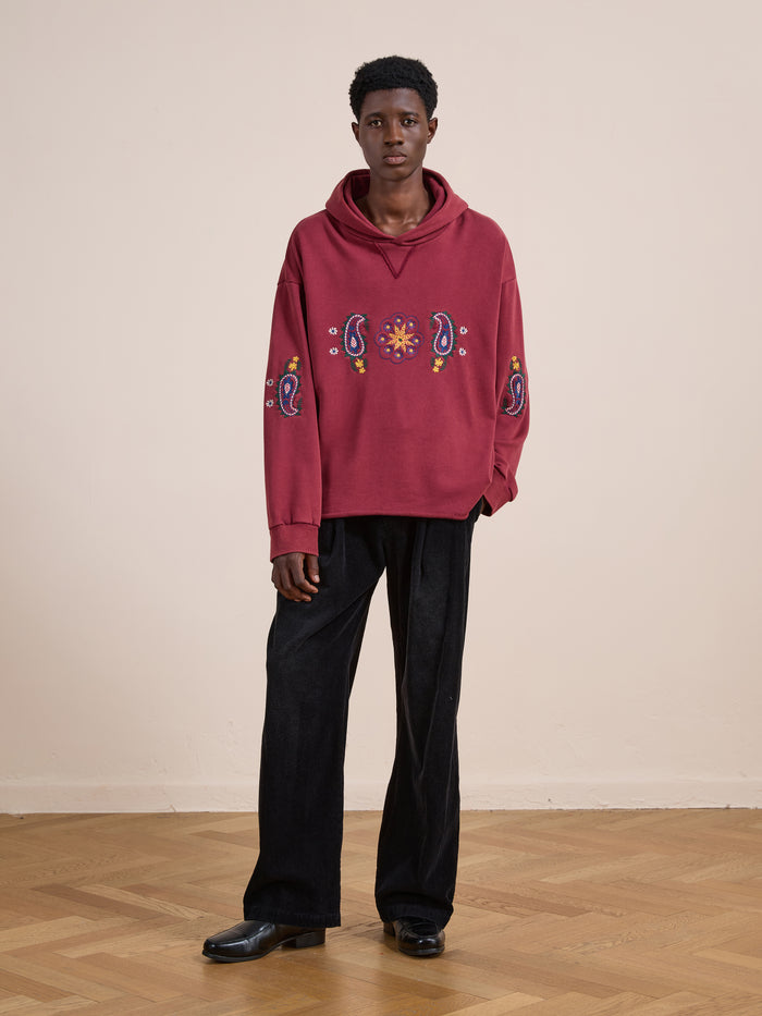 Indoors, a person wears the FOUND Embroidered Kantha Paisley Hoodie in maroon cotton fleece with paisley embroidery, paired with unisex relaxed fit black wide-leg pants and black shoes, set against a light wall and wooden floor.
