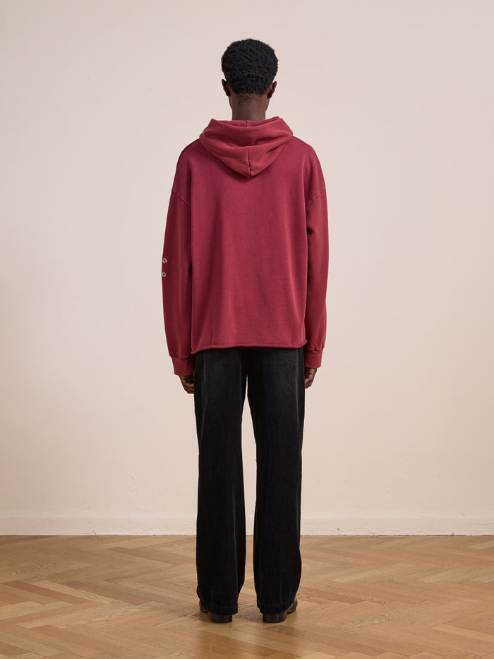 A person stands indoors with their back turned, wearing the FOUND Embroidered Kantha Paisley Hoodie in maroon cotton fleece with a unisex relaxed fit, paired with black pants on a wooden floor against a plain beige wall.