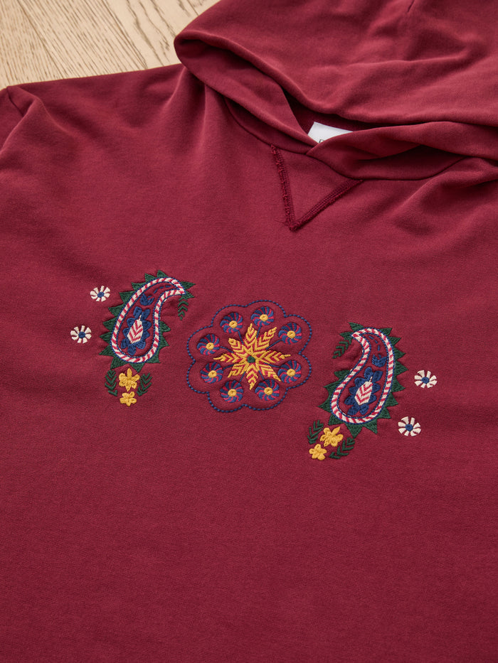 The FOUND Embroidered Kantha Paisley Hoodie is a maroon unisex cotton fleece with vibrant paisley and floral embroidery on the front, shown laid flat on a wooden surface.