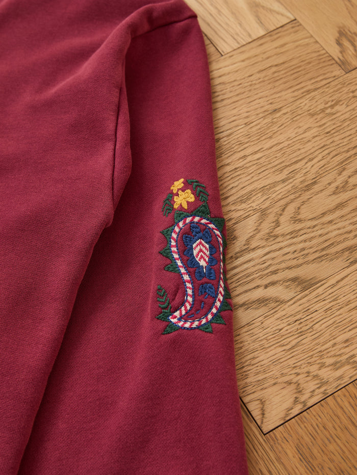 The FOUND Embroidered Kantha Paisley Hoodie is a maroon unisex relaxed fit cotton fleece with Kantha-inspired paisley embroidery and yellow flowers on the sleeve, displayed on a light wood floor.