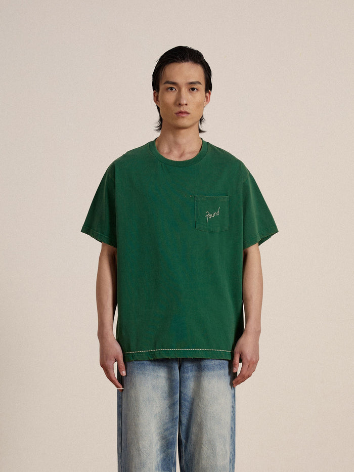 A man wearing a green Found embroidered logo tee and jeans with distressing.