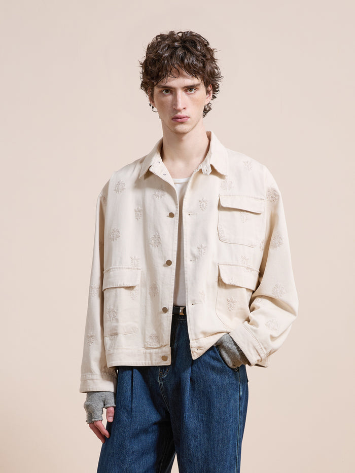 A young person with curly hair wears the Found Embroidered Motif Jacket in a unisex relaxed fit, featuring two front pockets, over blue jeans while standing against a plain beige background.