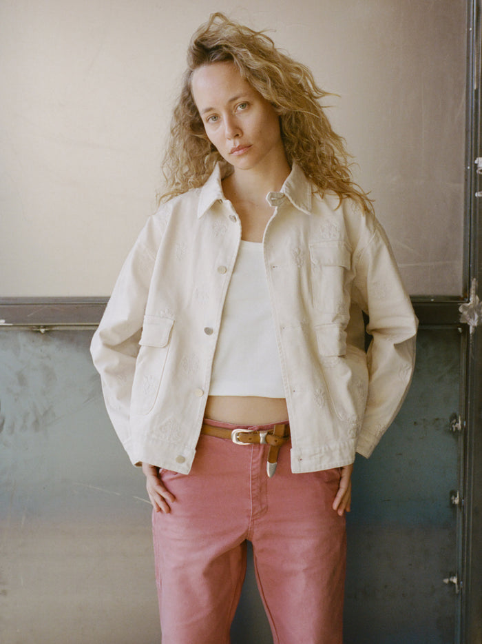 Woman with wavy hair wearing the Found Embroidered Motif Jacket over a white top and pink pants, standing against a metallic background with hands in her pockets.