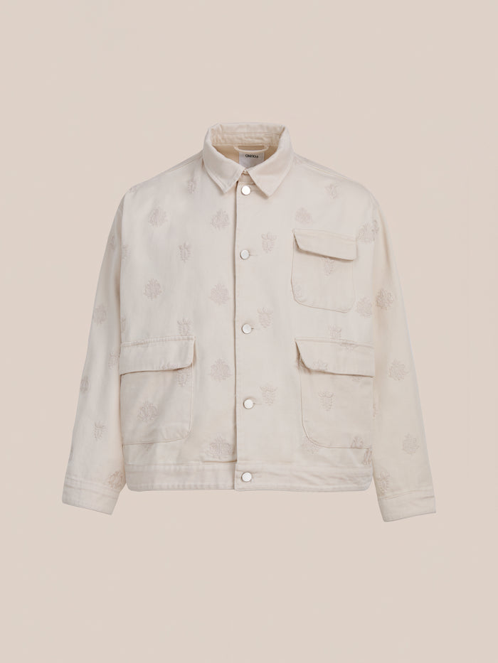 The Found Embroidered Motif Jacket is a cream-colored unisex vintage chore jacket with three front pockets and subtle embroidery, shown on a plain beige background.