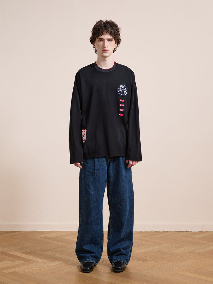 Indoors on a wooden floor, a person wears FOUND’s Embroidered Patches LS Tee in vintage washed black with distressed details and "END CLUB" text, paired with wide blue jeans against a beige wall.