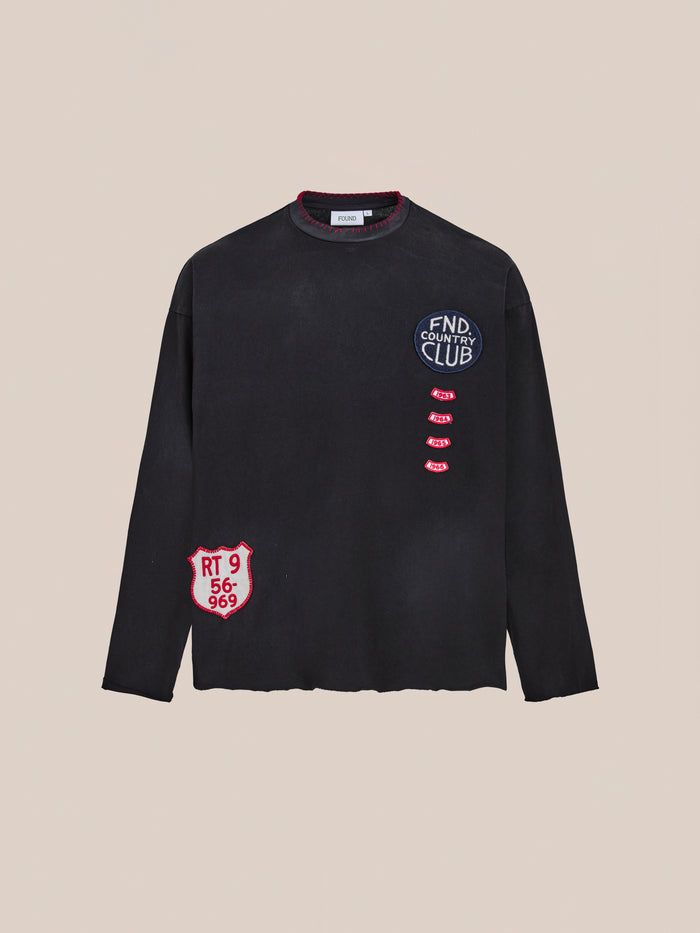 The FOUND Embroidered Patches LS Tee is a vintage washed black long-sleeve shirt featuring distressed details and red and white embroidered patches, including "FND COUNTRY CLUB" on the chest and "RT 9 56-969" near the hem.