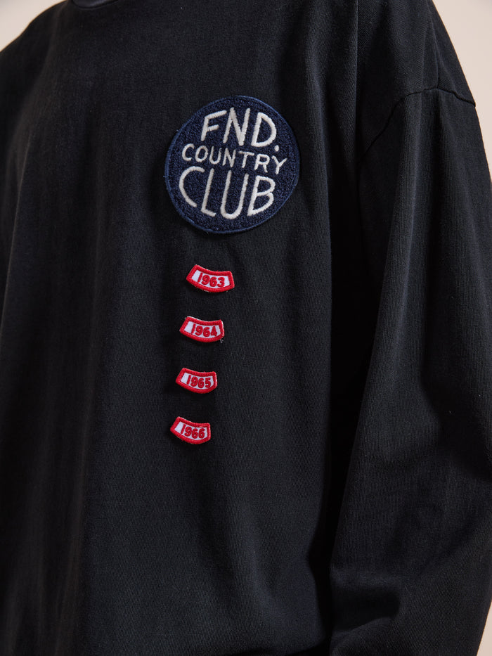 The FOUND Embroidered Patches LS Tee is a vintage washed black shirt with a "FND Country Club" patch and four small red patches below, each embroidered with the years 1963, 1964, 1965, and 1966.