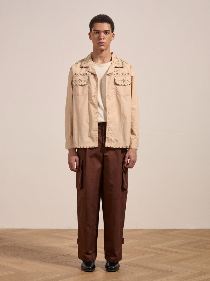 A person is standing indoors, wearing the FOUND Embroidered Western Button Up in beige, a white T-shirt, and brown pants with large pockets. The floor is wooden, and the background wall is plain.