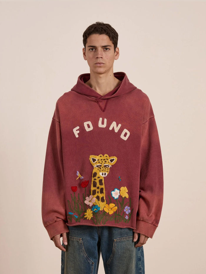 Someone stands against a neutral background wearing the FOUND Embroidered Wildlife Hoodie, which features embroidered flowers, butterflies, a bee, and "FOUND" on the front.