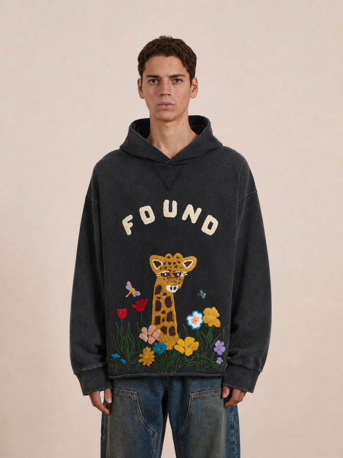 A person wearing the FOUND Embroidered Wildlife Hoodie, featuring a dark French-terry cotton fabric with “FOUND” text, a giraffe, flowers, and butterflies embroidered on the front.